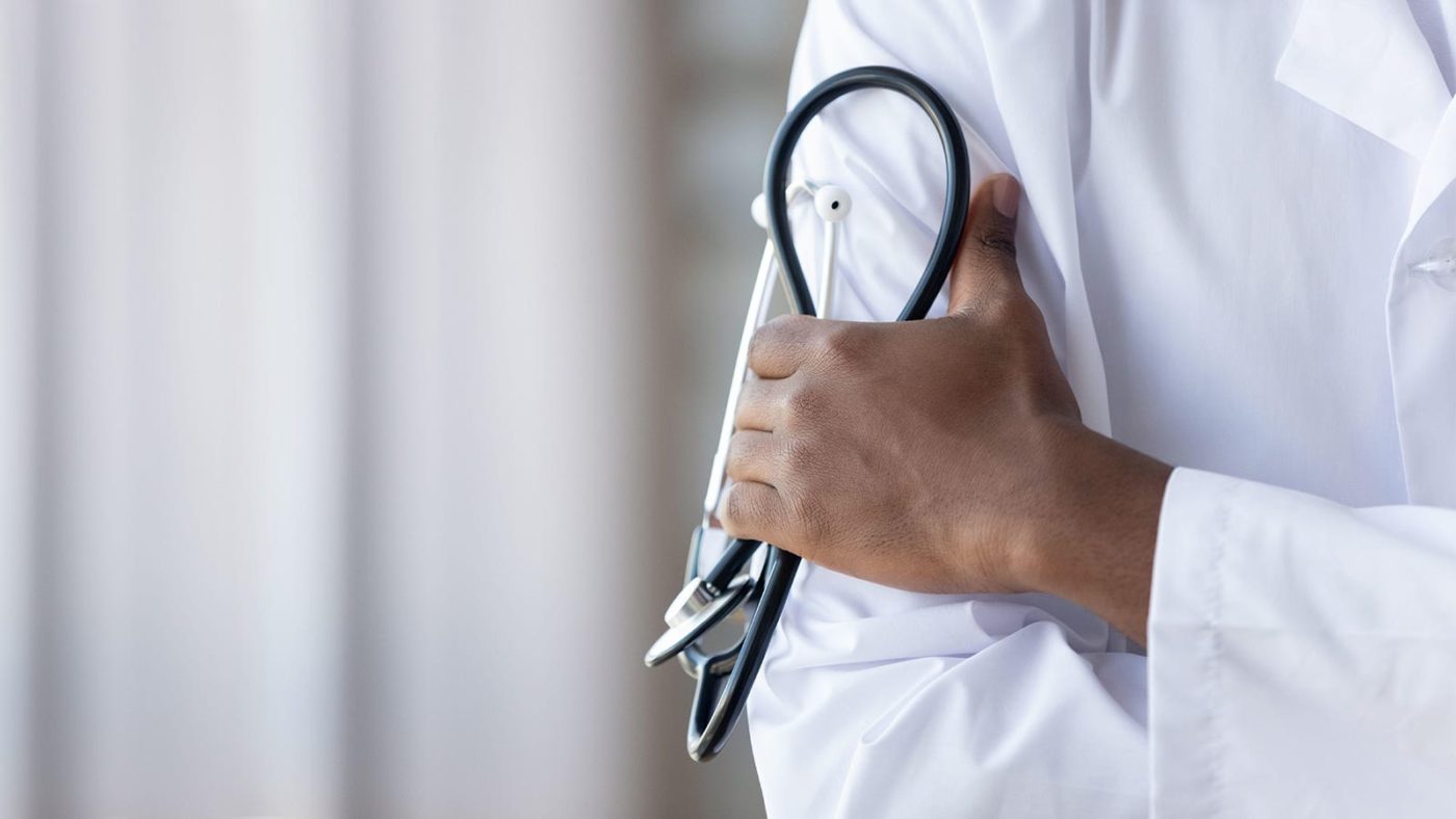 A Black hand clutches a stethoscope on the sleeve of an arm in a white coat.
