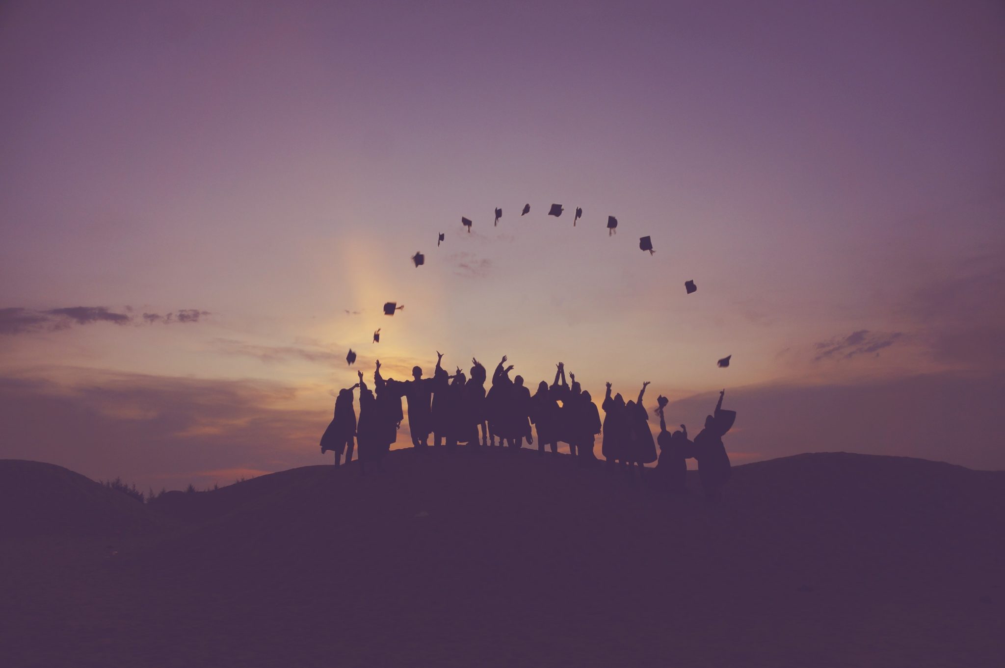 Photo of students throwing graduation caps into the air against a backdrop of the setting sun