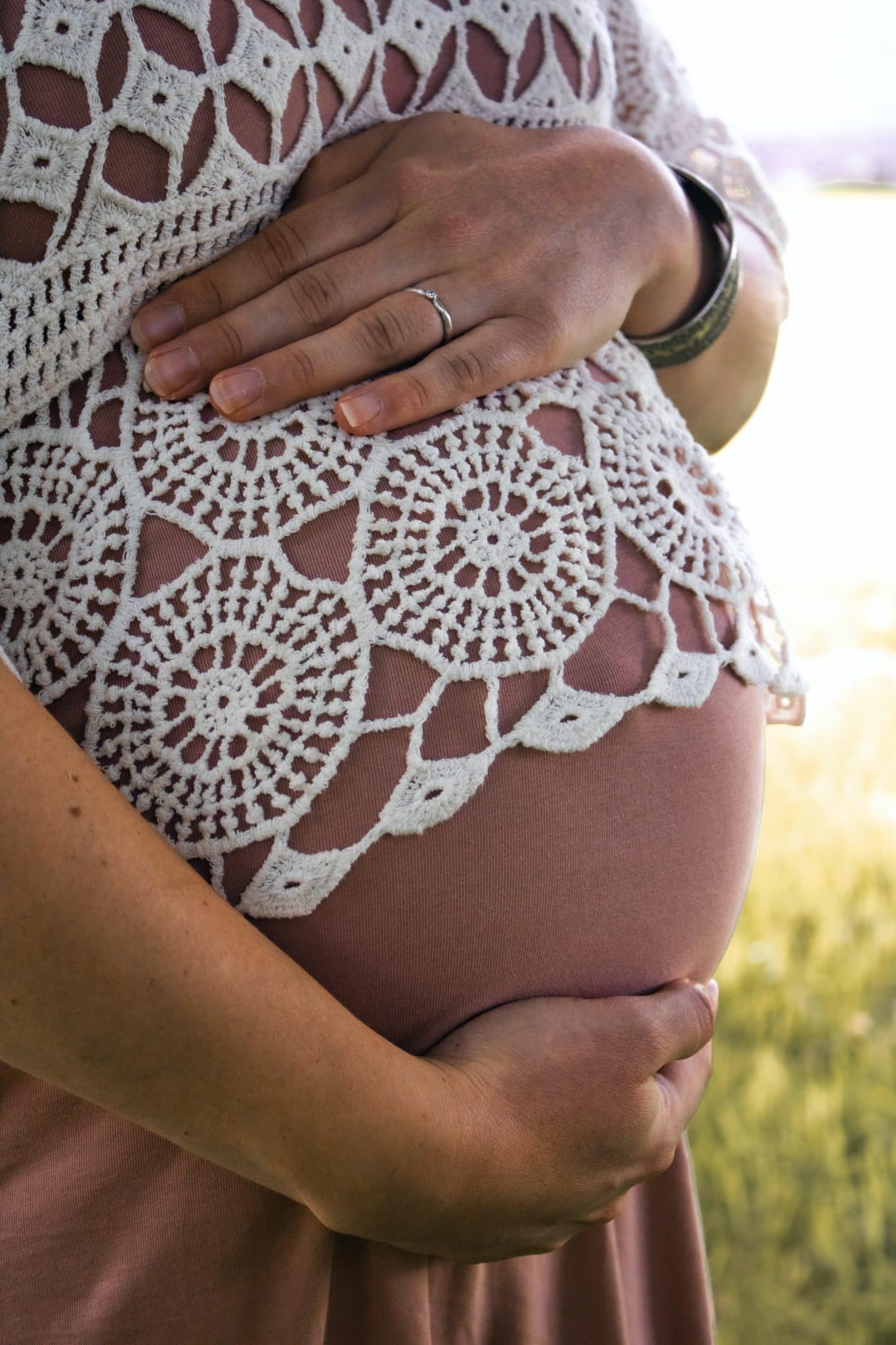 Photo of a pregnant woman wearing a white lace top, holding her belly with both hands.
