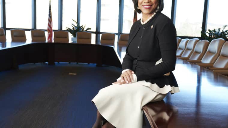 Photo of Dr. Valerie Montgomery Rice leaning against a conference table with windows and an American flag in the background