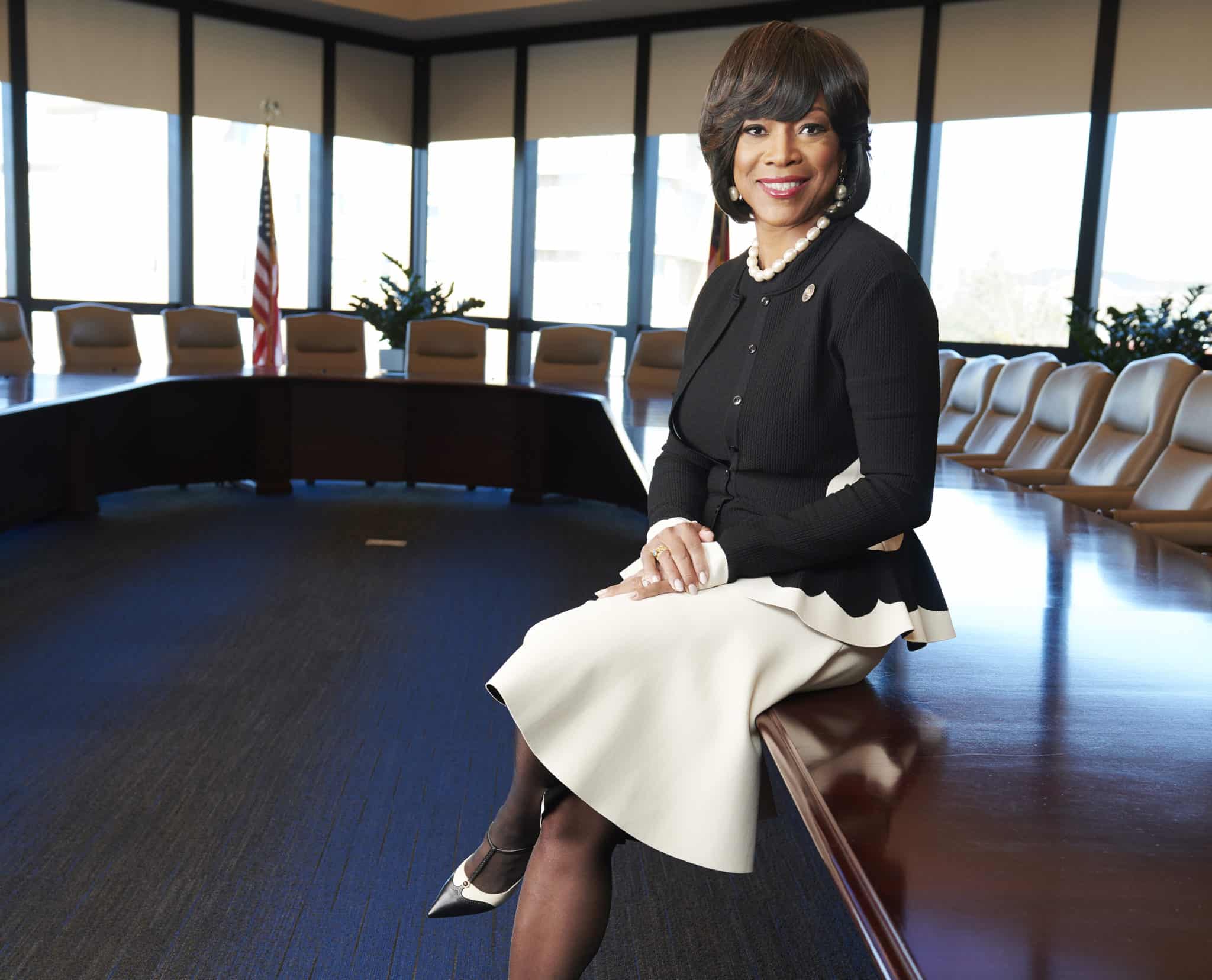 Photo of Dr. Valerie Montgomery Rice leaning against a conference table with windows and an American flag in the background