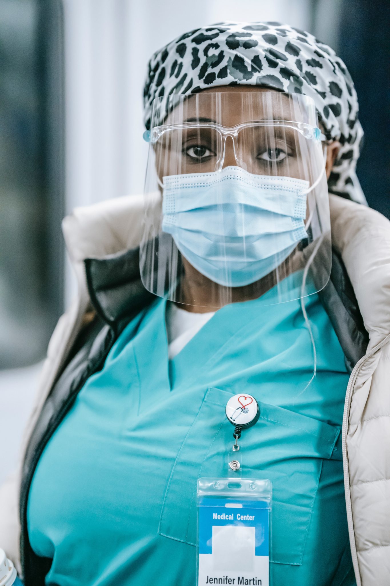 A black health care worker wearing blue scrubs, a coat, a face mask and face shield looks into the camera