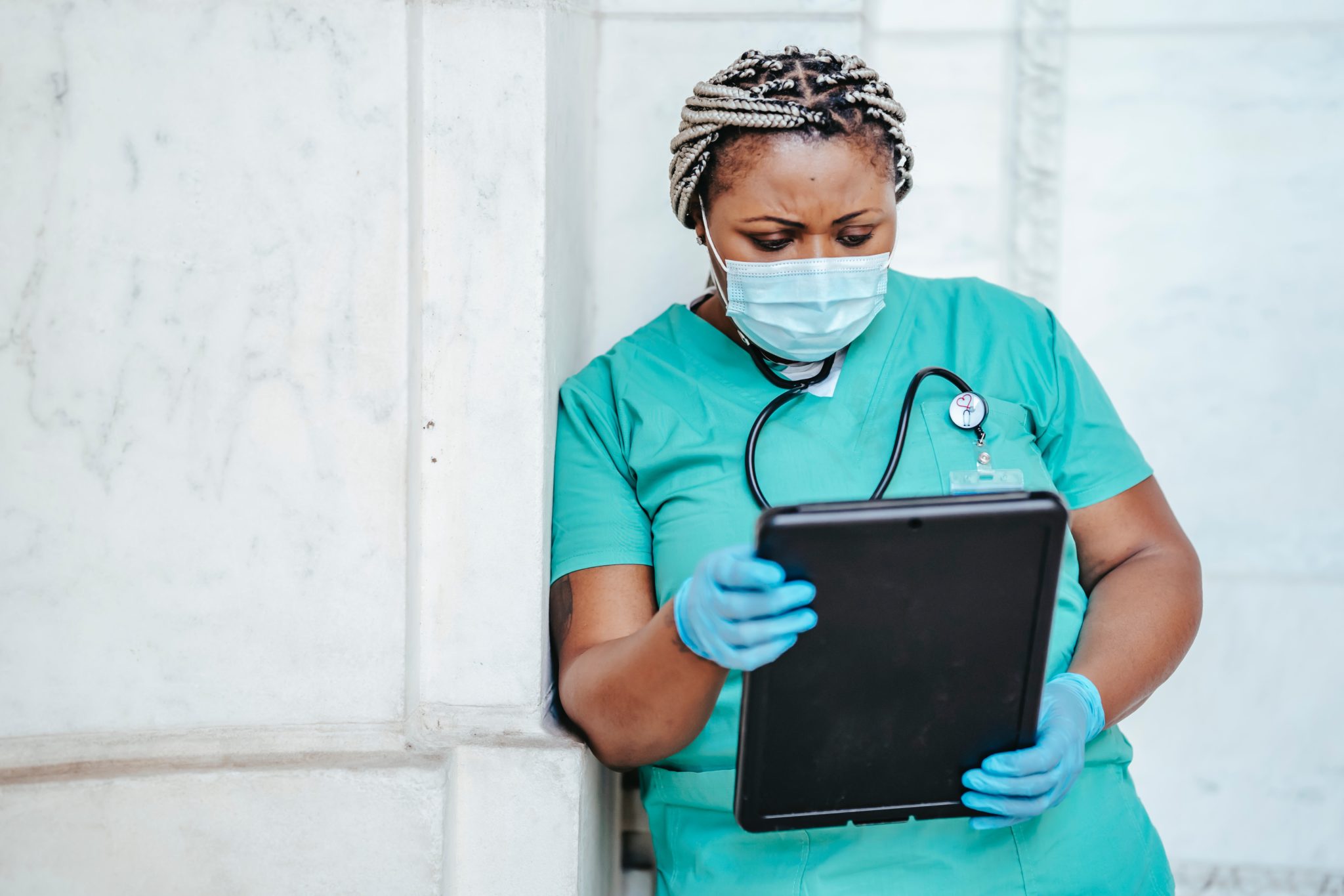 A Black doctor or nurse wearing a stethoscope looks at a tablet that they are holding while wearing gloves.