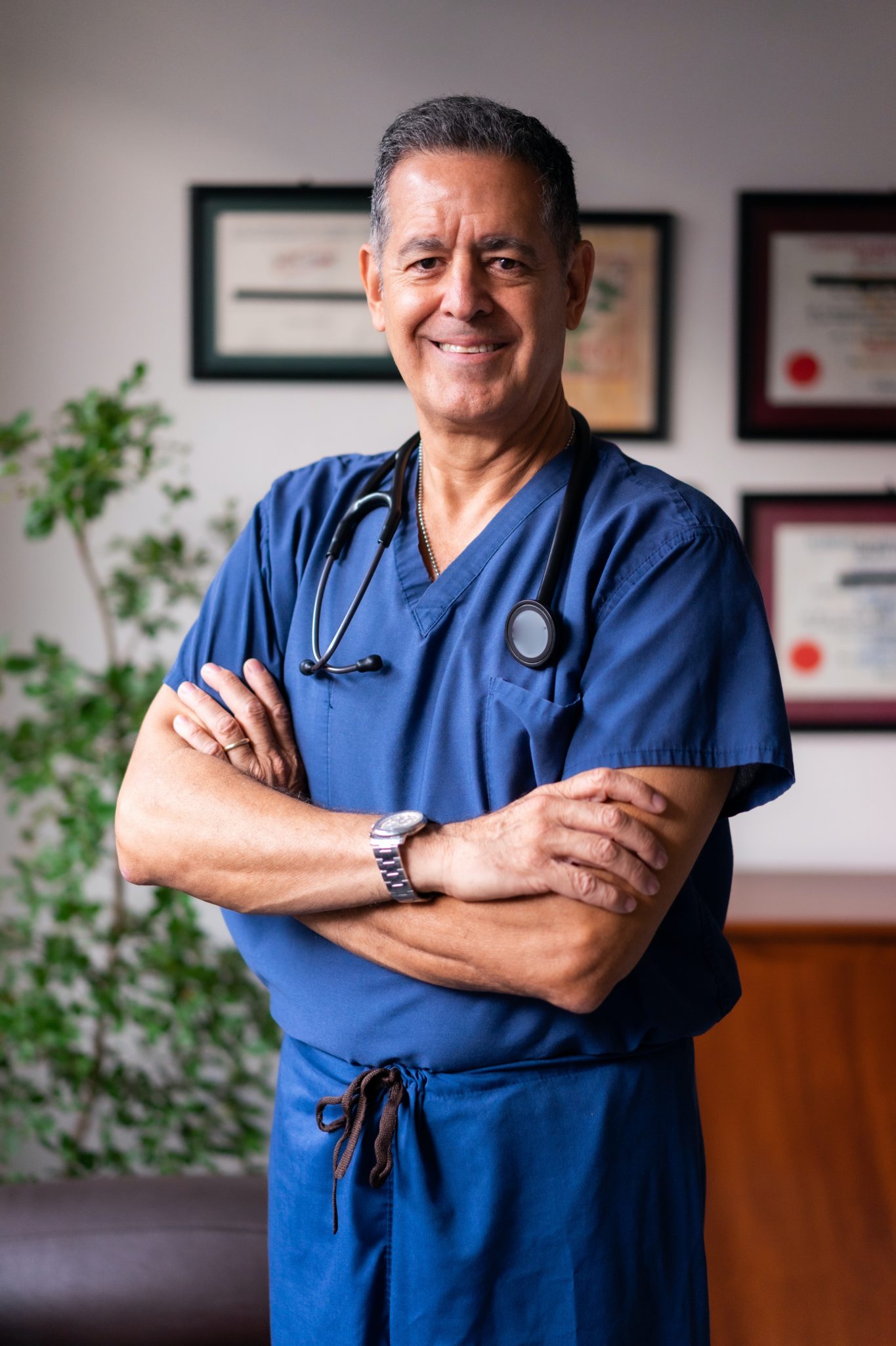 A Latine doctor stands with is arms crossed in blue scrubs smiling to the camera