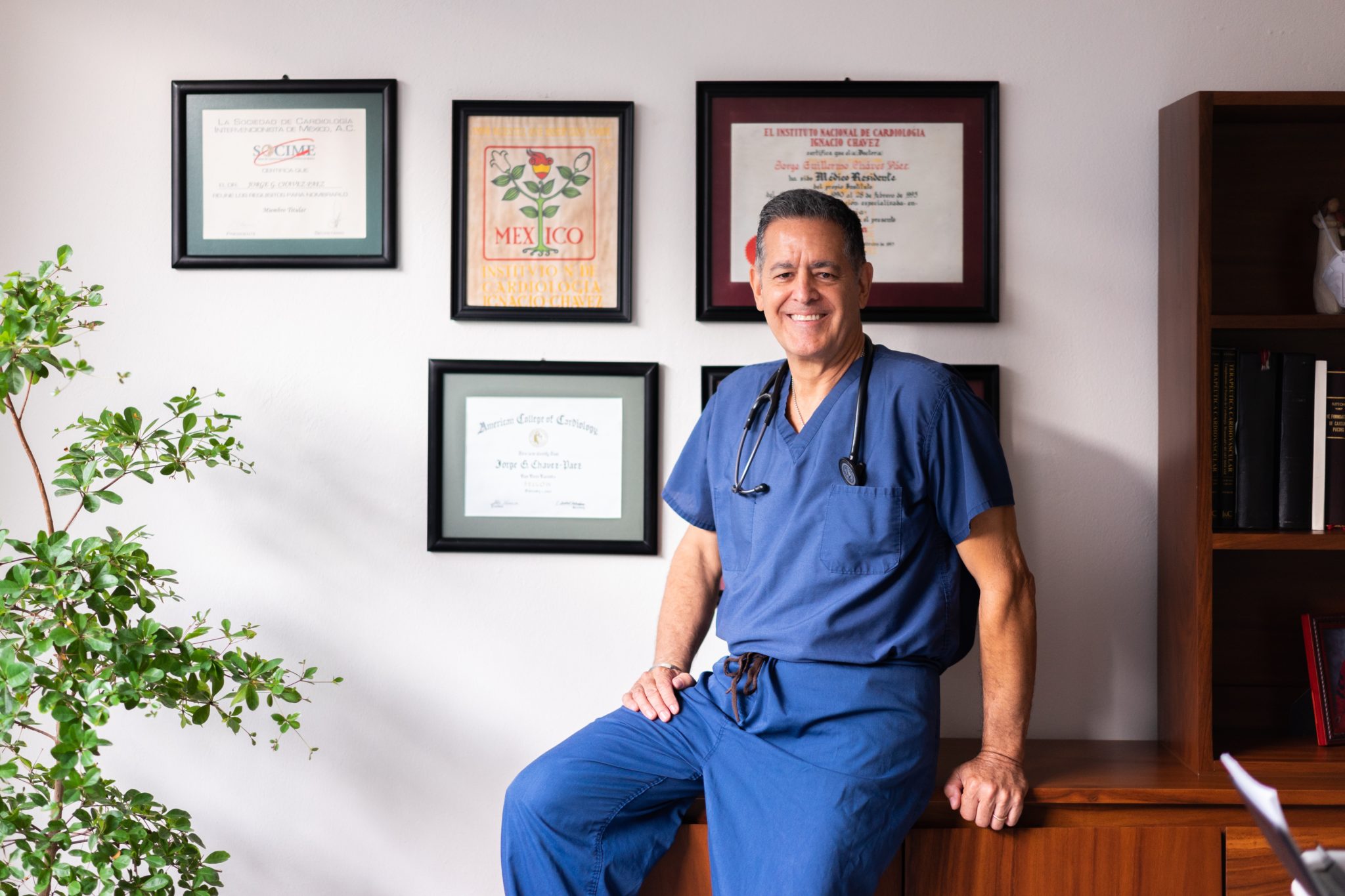 A Latine doctor sits on a cabinet in an office that has a degree on the wall and a poster reading Mexico