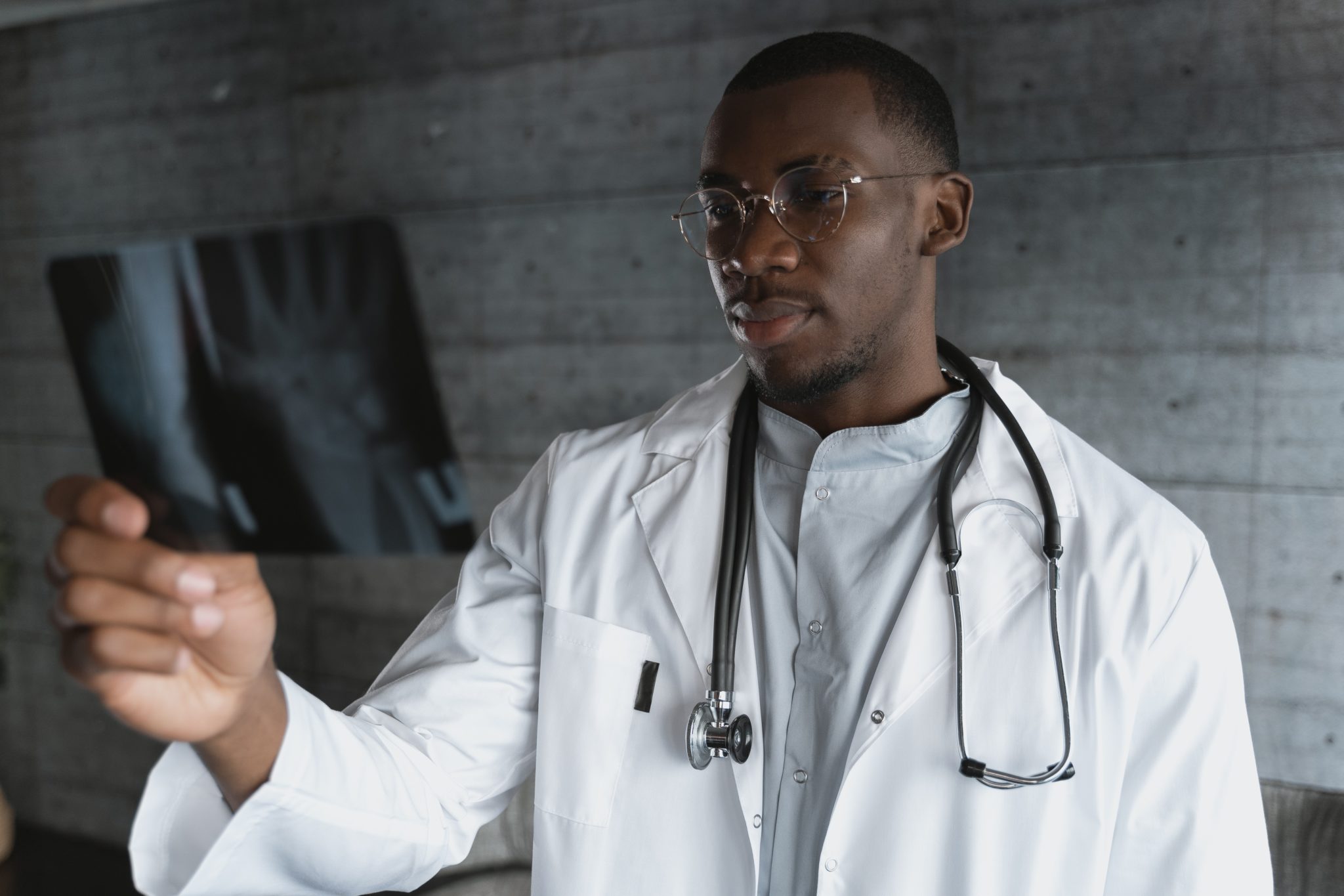 A black doctor wearing a white coat and glasses looks at an xray