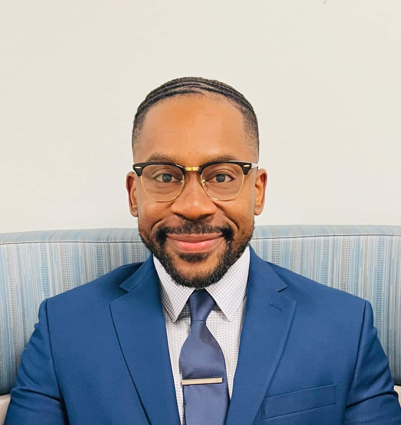 Photo of Dr. Michael Enechukwu wearing a blue blazer and tie with a white shirt. He is wearing glasses.