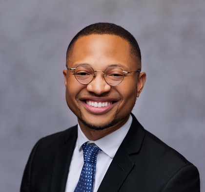 Photo of Jeremiah Wittington smiling into the camera. He is wearing a black blazer and blue tie and glasses