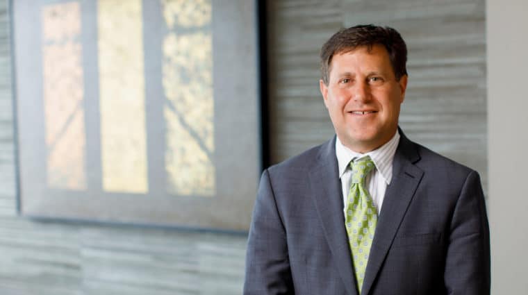 Photo of Scott Becker wearing a gray jacket, white shirt, and green tie. He is standing in front of a wall with a shaded window in the background