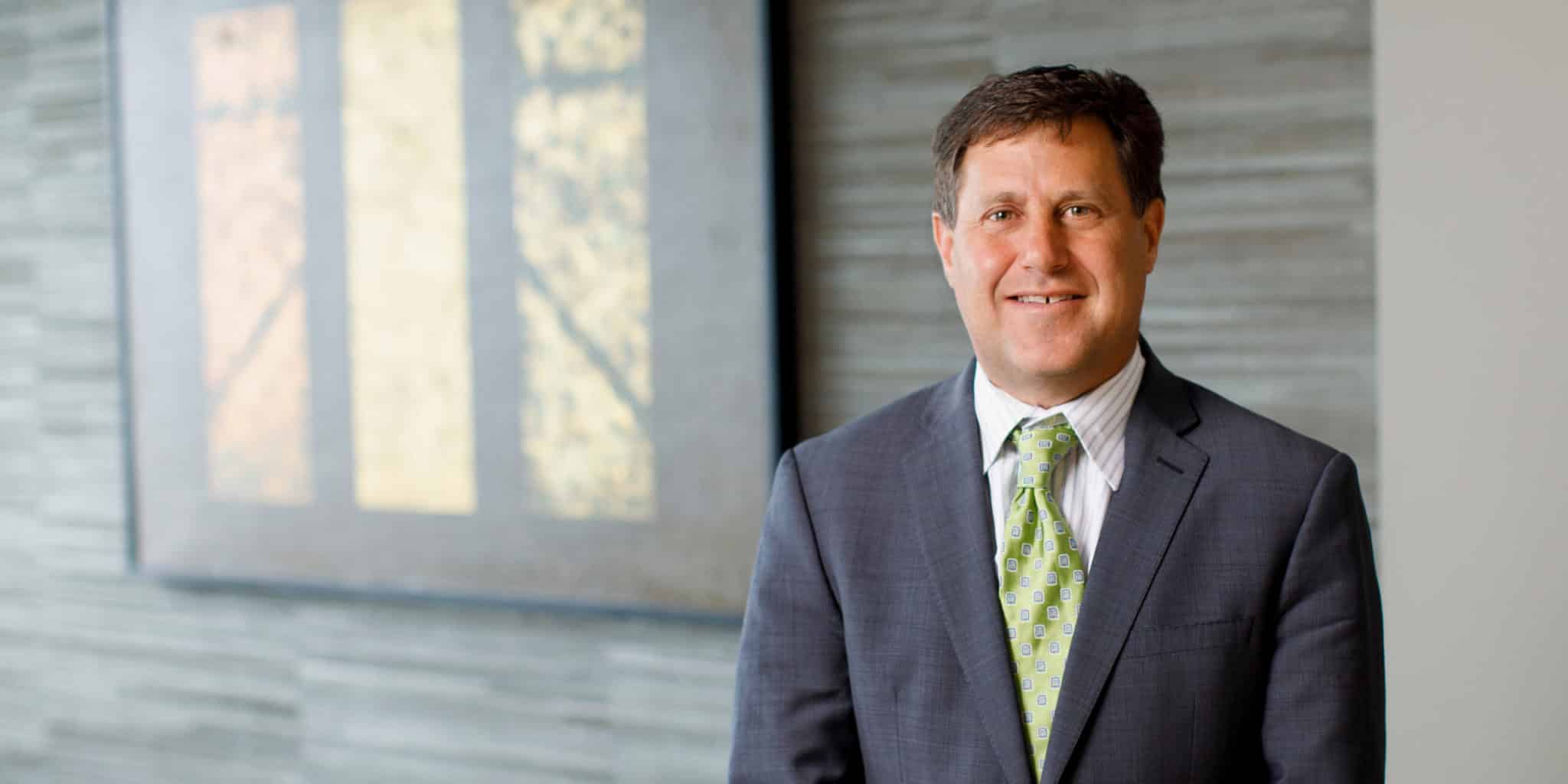 Scott Becker Photo of Scott Becker wearing a gray jacket, white shirt, and green tie. He is standing in front of a wall with a shaded window in the background