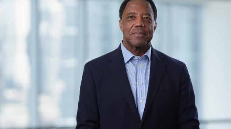 Photo of Lloyd Dean wearing a black jacket and blue shirt, standing in front of a large window