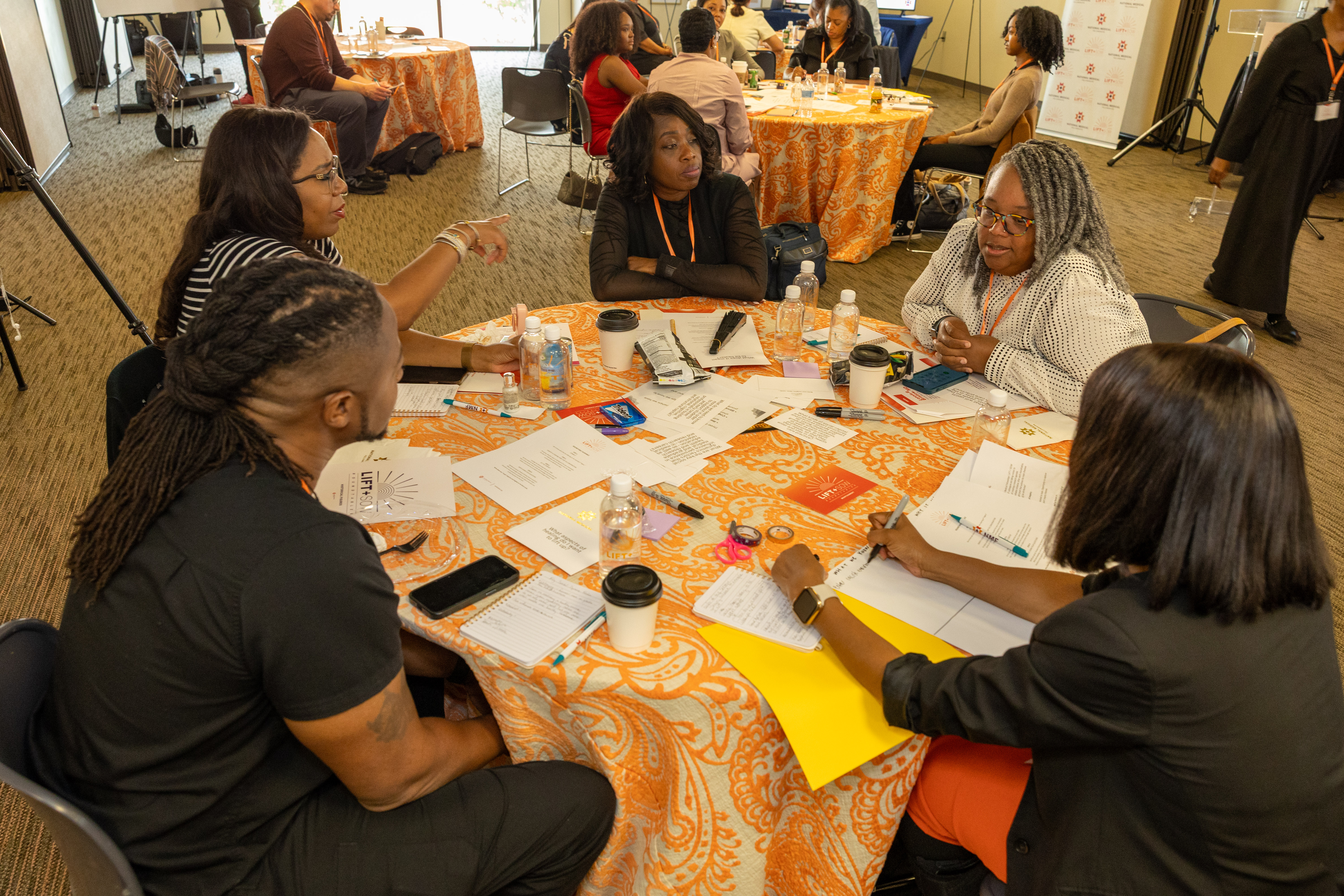 A group of roundtable participants are speaking to each other. They are at a round table with an orange table cloth.