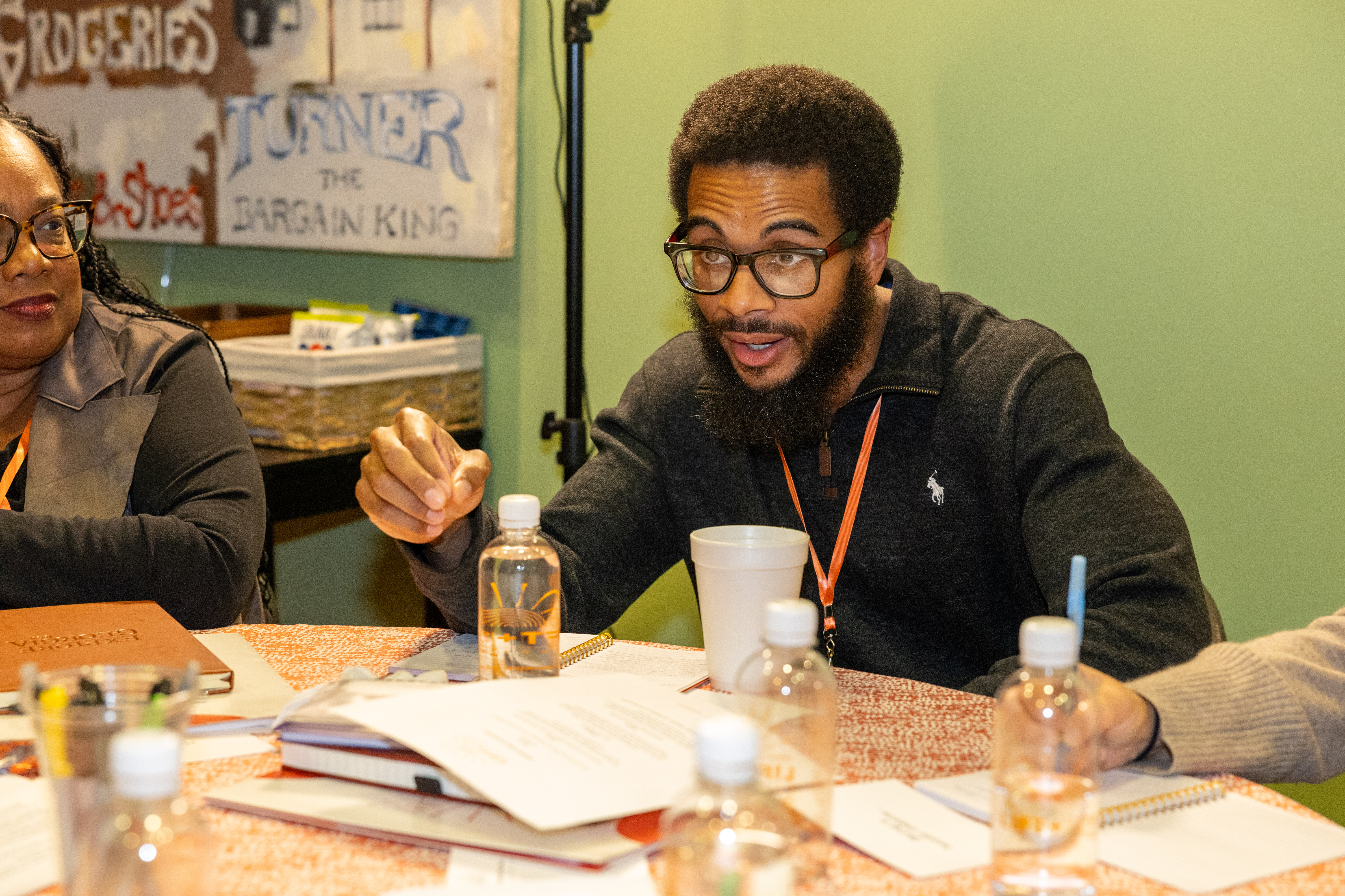 A man dressed in a black sweater and wearing glasses gestures as he speaks, sitting at a round table