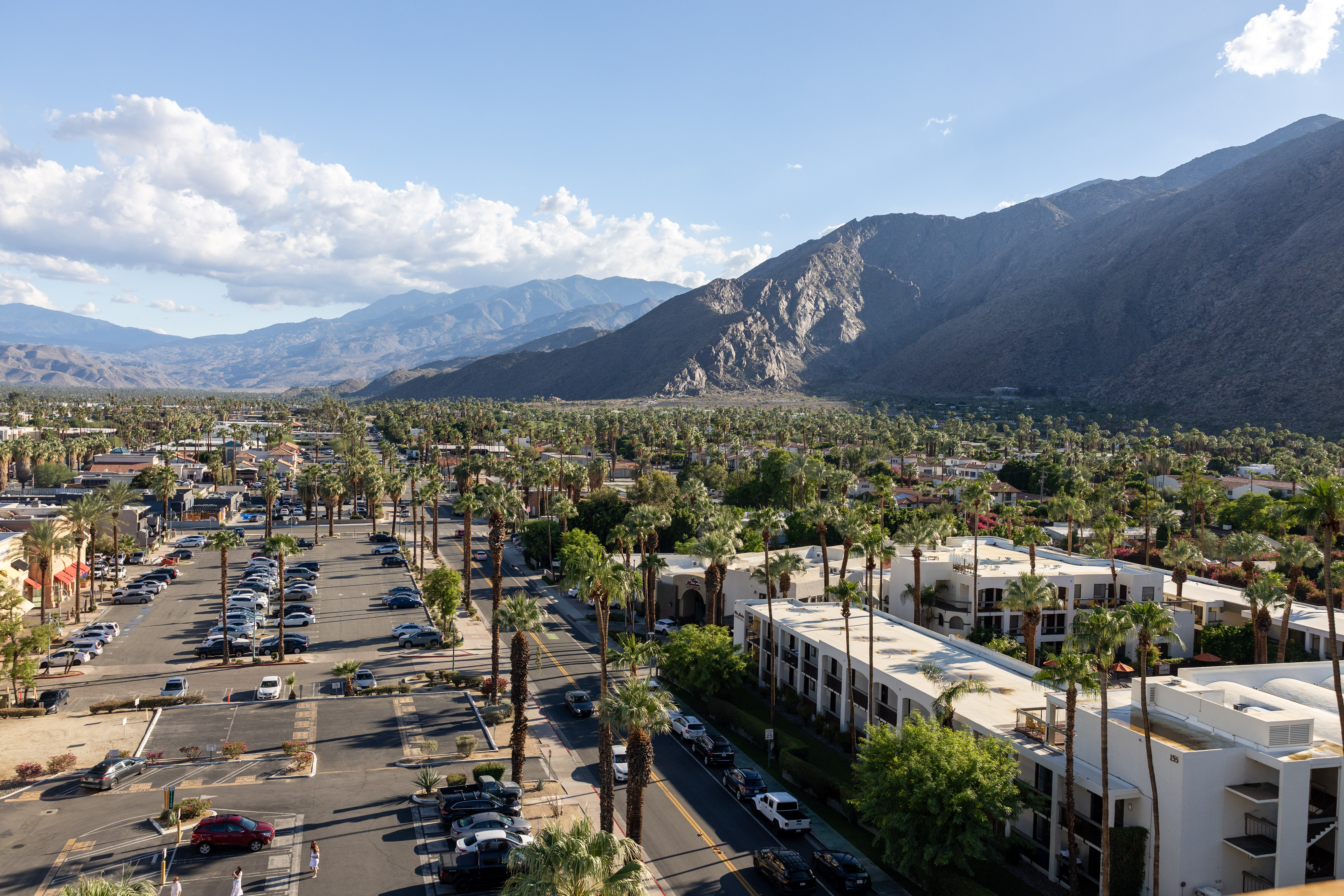 A photo of Palms Springs, featuring a downtown with roads and buildings, and tall mountains behind, with a blue sky and white clouds