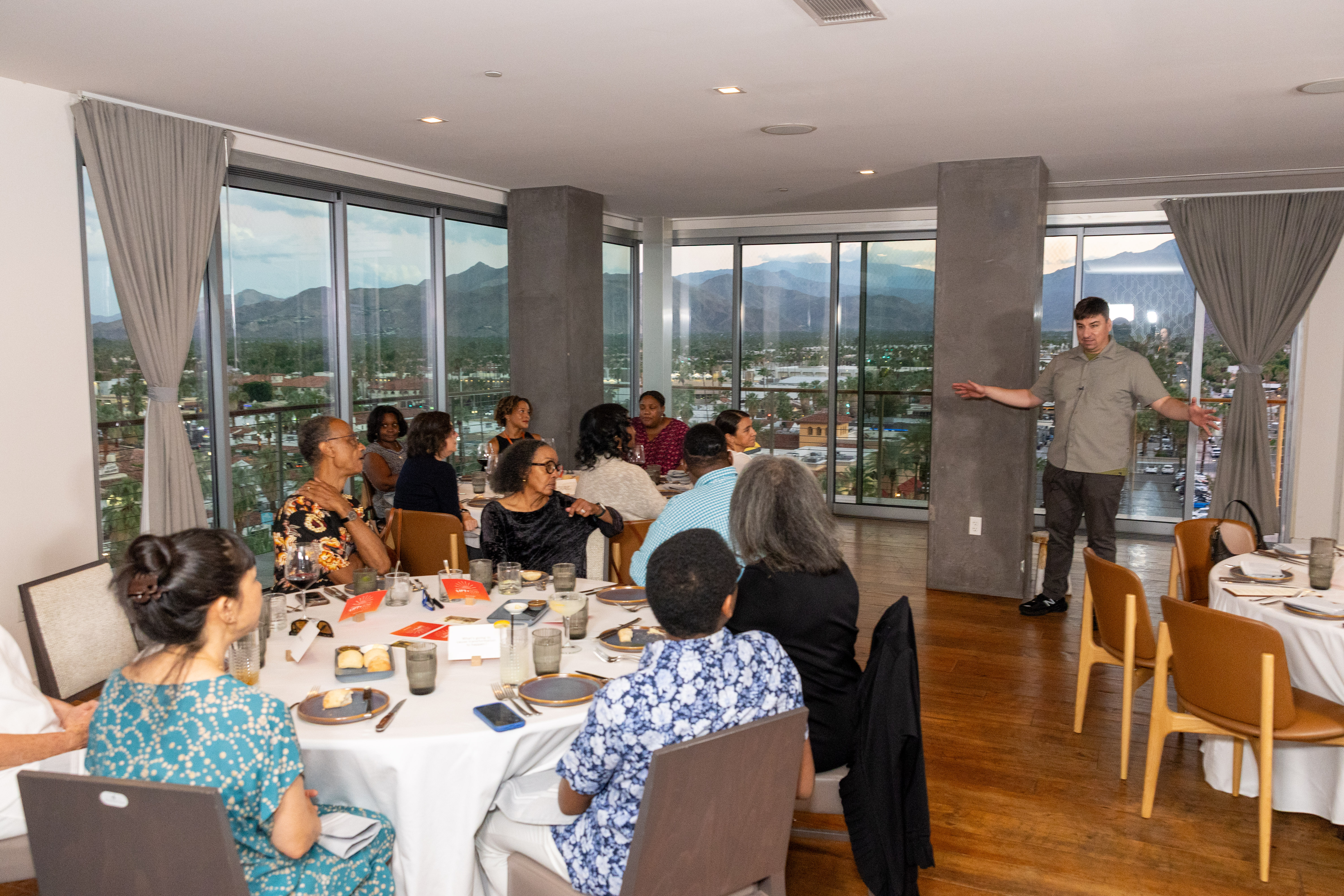 Several tables of people are seated listening to a speaker make a presentation