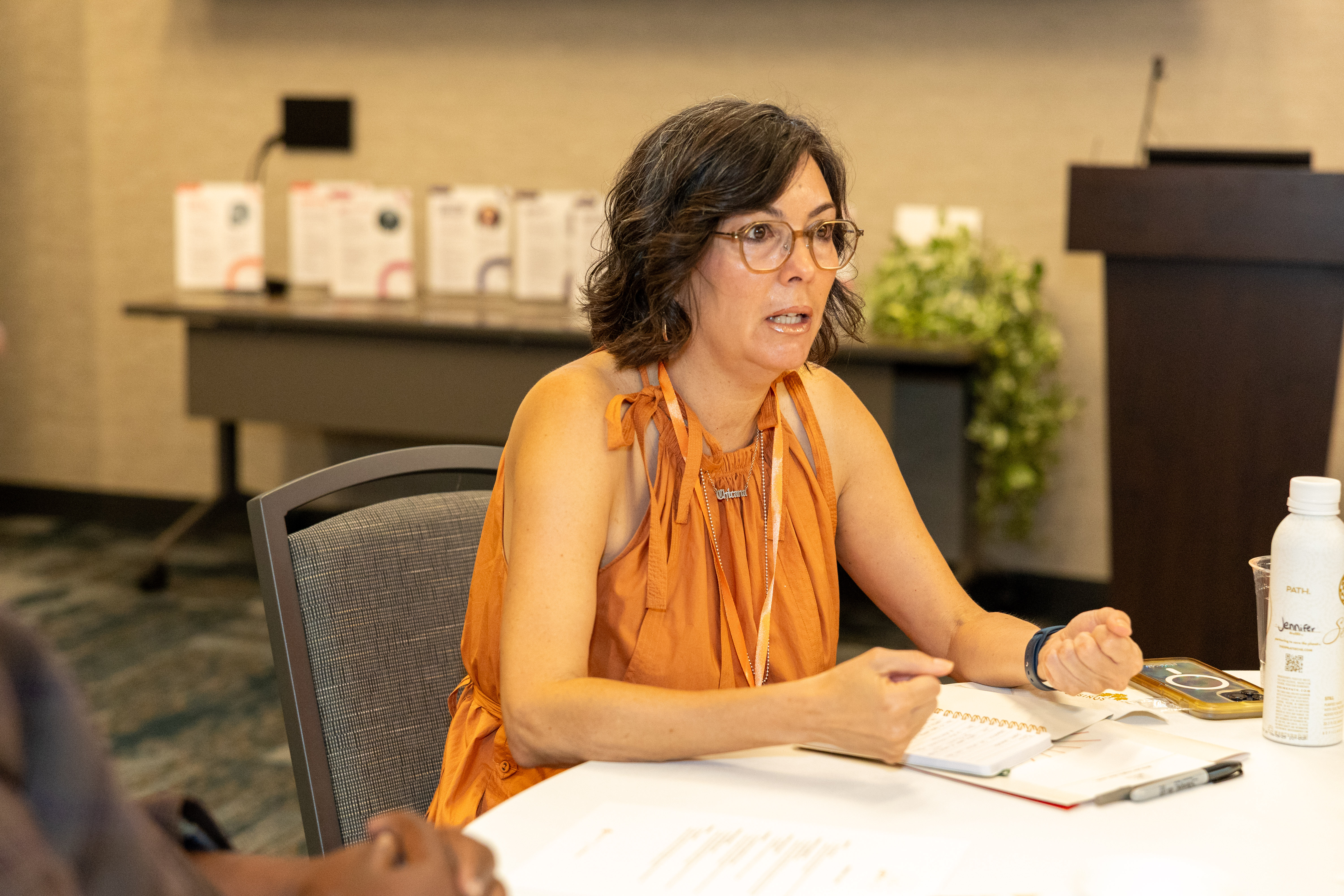 A woman in a sleeveless orange blouse wearing glasses is seated at a table and participating in a discussion.