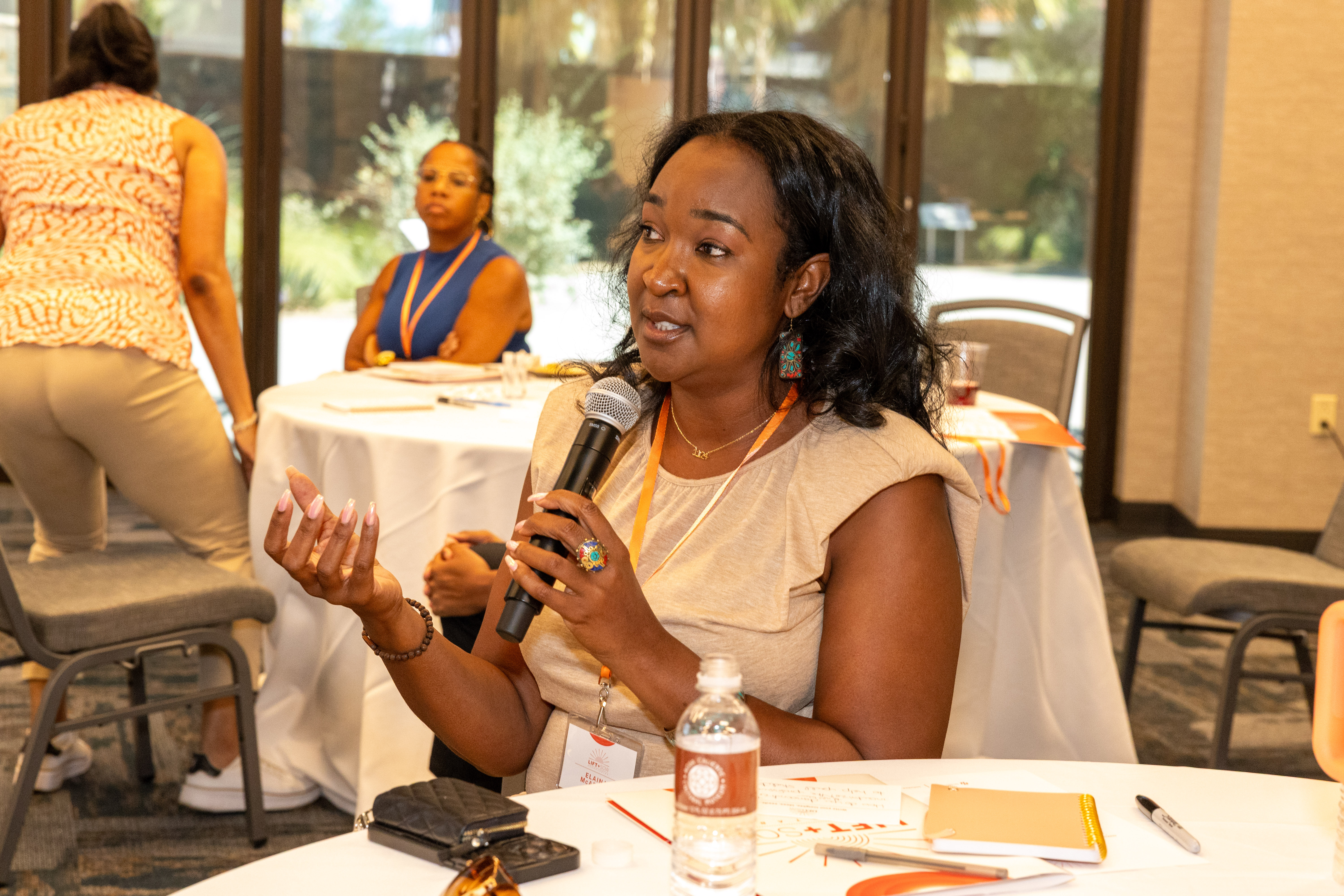 A woman in a white blouse is gesturing with her hands as she is speaking into a microphone. She wears a white blouse