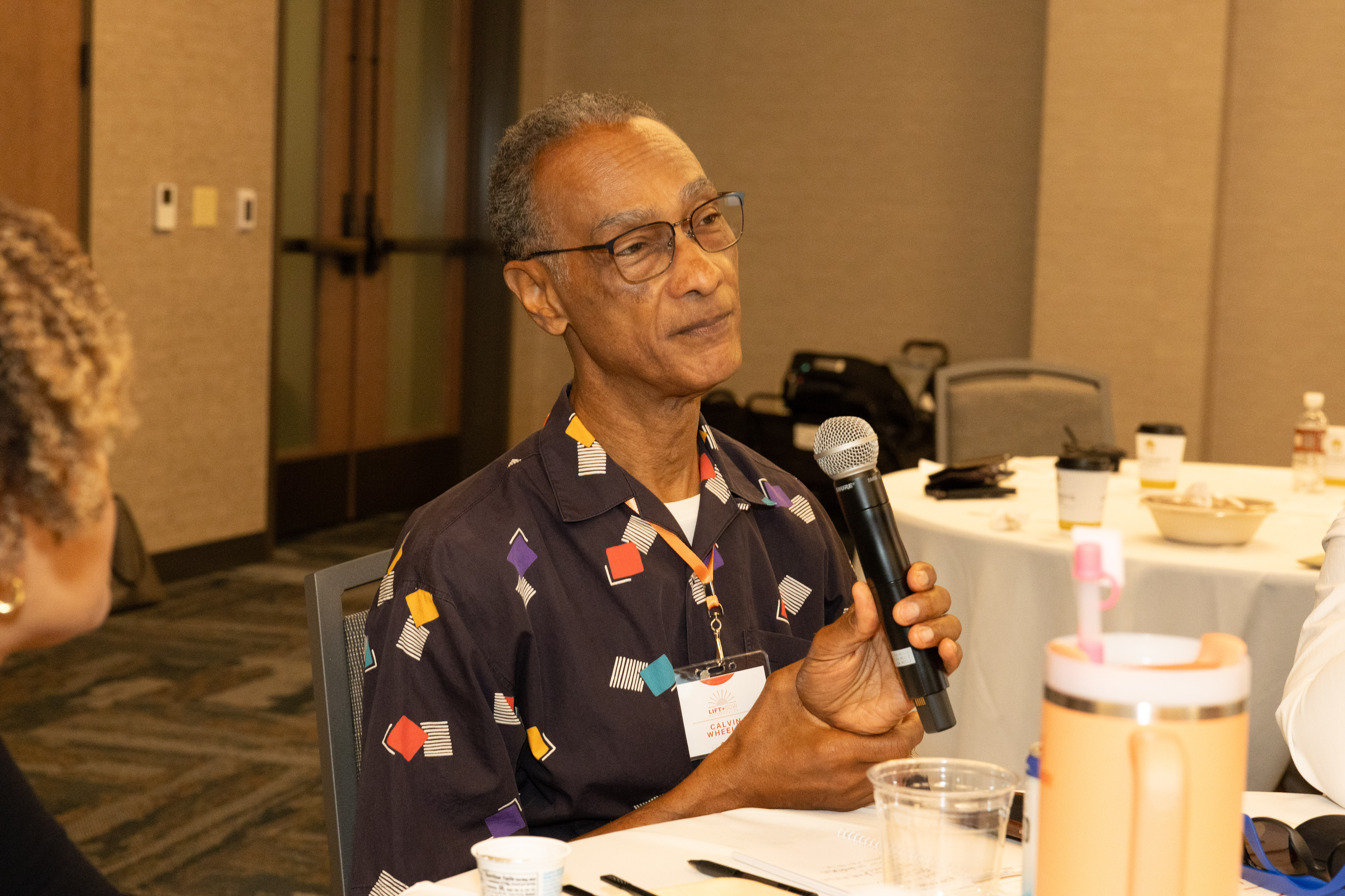 A photo of Dr. Calvin Wheeler speaking into a microphone. He is wearing glasses and a brown shirt with multicolored blocks on it.