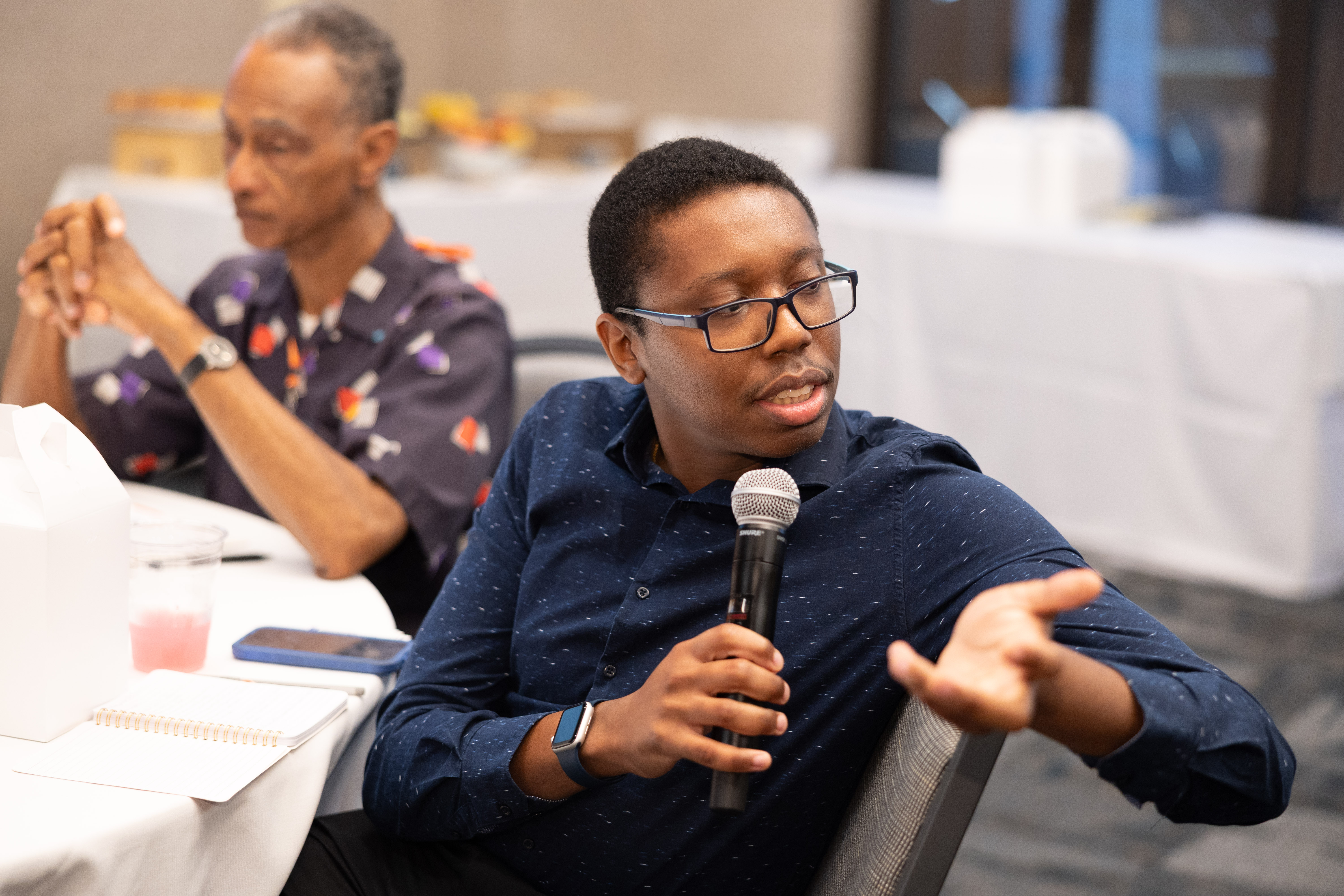 A young man seated at a table speaks into the mic. He is wearing a blue shirt and glasses.