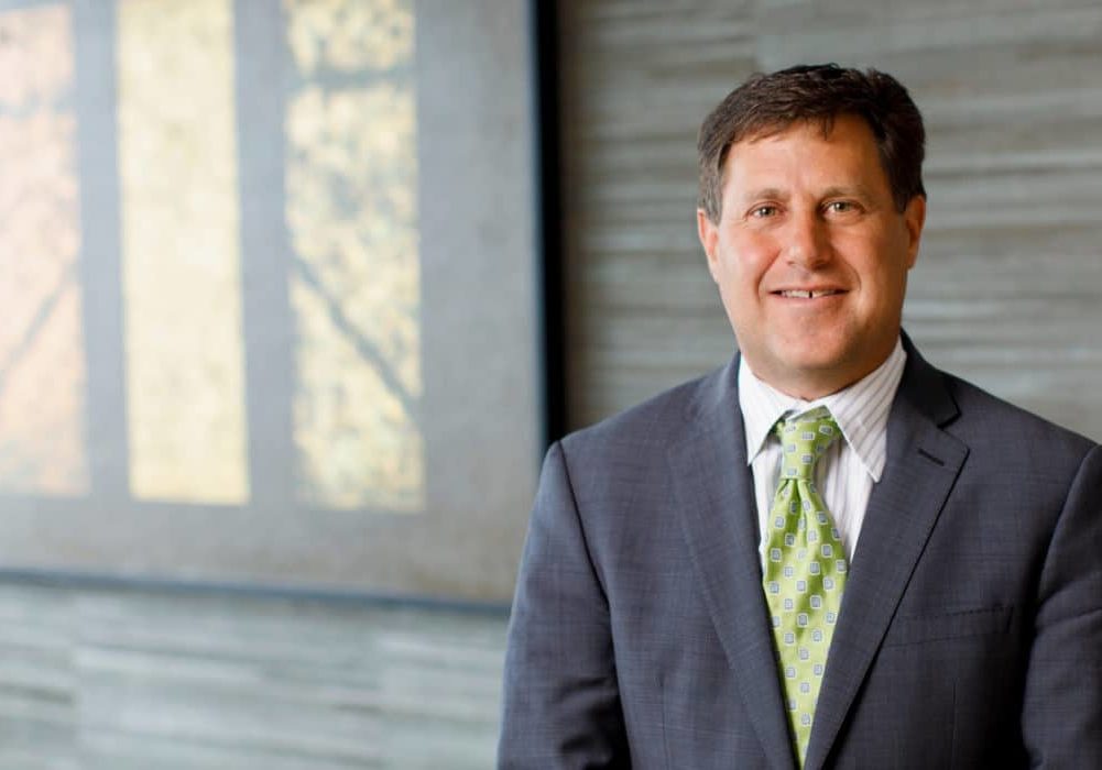 Photo of Scott Becker wearing a gray jacket, white shirt, and green tie. He is standing in front of a wall with a shaded window in the background