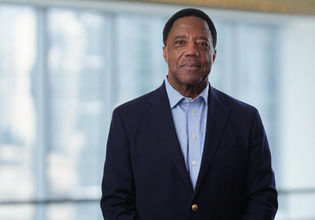Photo of Lloyd Dean wearing a black jacket and blue shirt, standing in front of a large window