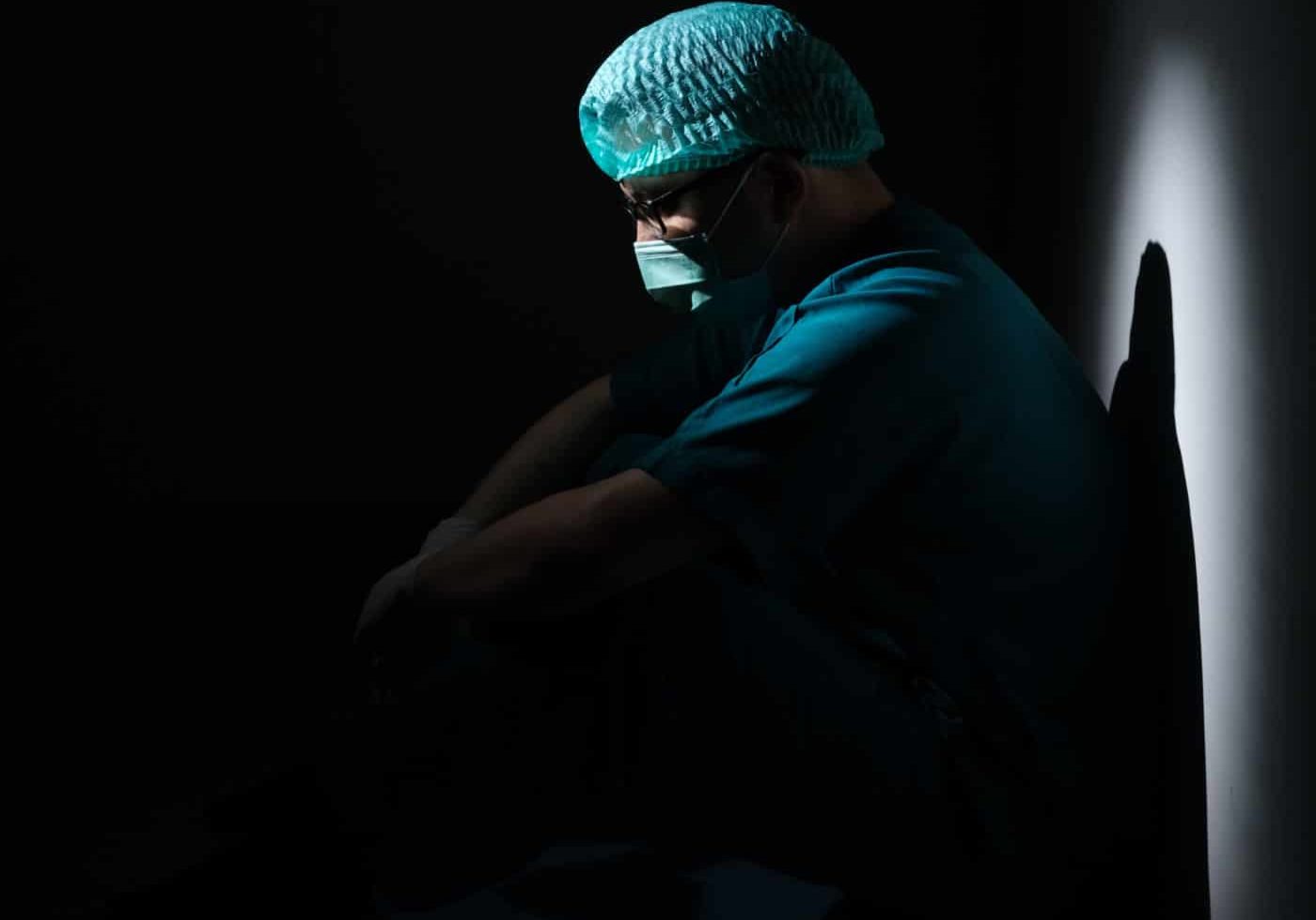 Photo of an exhausted doctor in mask and scrubs sitting in a dark room
